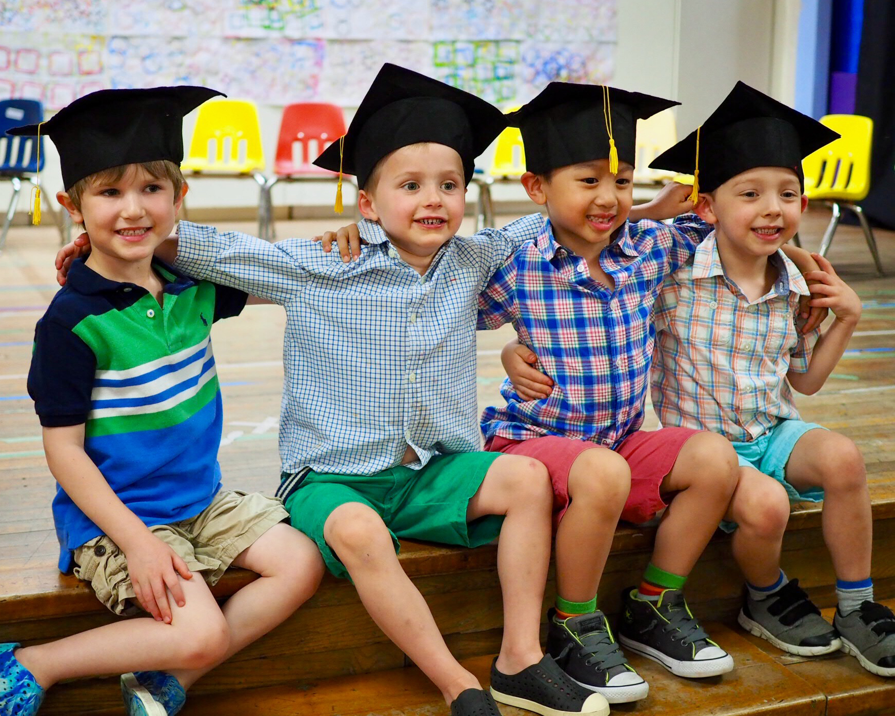 Students in Graduation Caps 4 young boys sitting on gymnasium stage with graduation caps on after their graduation ceremony